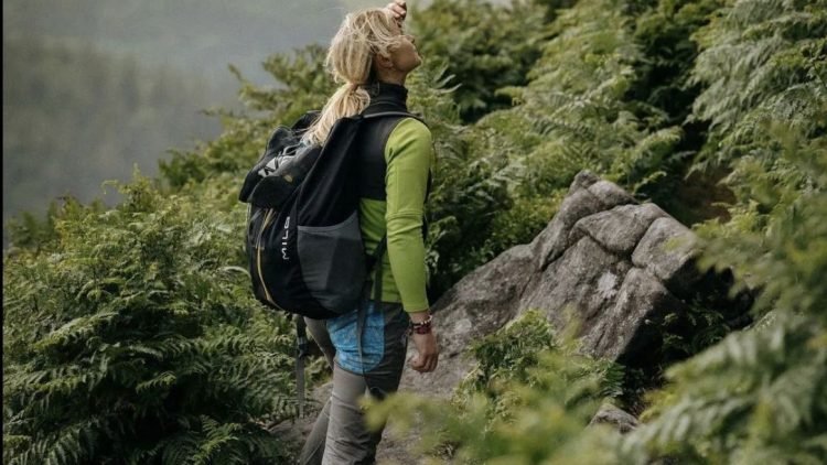 Mujer con mochila de montaña negra caminando por sendero rodeado de vegetación densa en zona montañosa.
