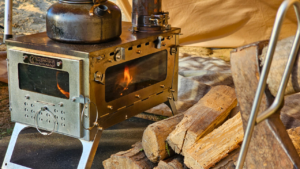 Cocina portátil de leña FASTFOLD S2W completa sobre fondo blanco mostrando estructura plegable de acero inoxidable, doble panel con ventanas de vidrio templado, chimenea modular y bolsa de transporte incluida